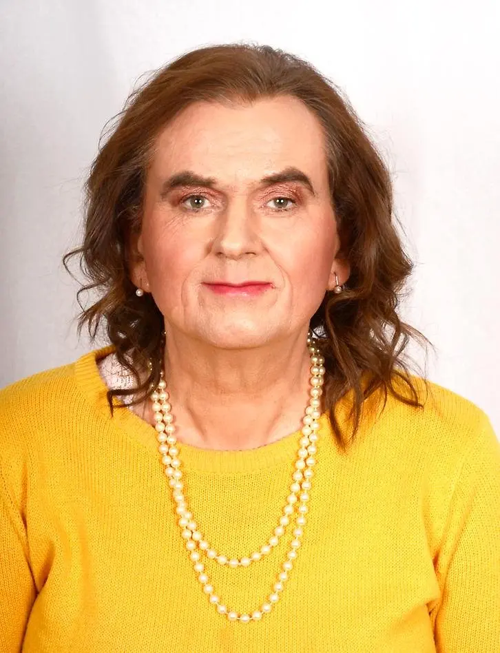 Kvinna med halvlångt brunt lockigt hår. Har på sig en varmgul tröja. Ovanpå den ett pärlhalsband.