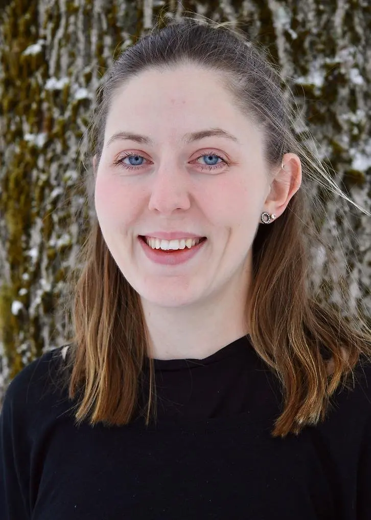 En leende kvinna med halvlångt brunt hår. Luggen är uppsatt bakåt. I vänster öra har hon örhängen.Hon har en svart tröja på sig. Bilden är tagen utomhus framför en trädstam.