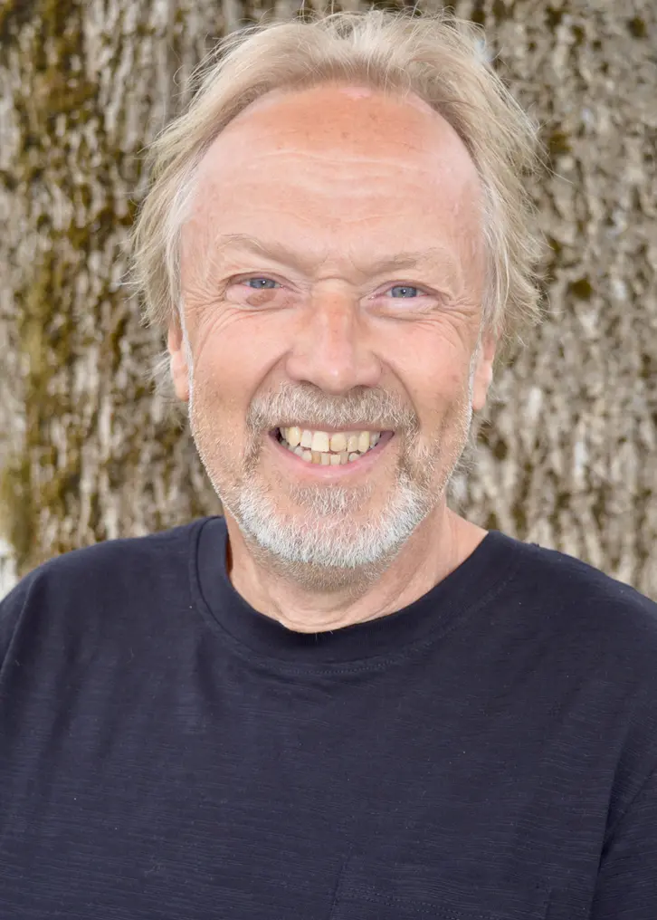 En leende man med kortklippt ljust hår. Han har kort skägg. På sig har han en svart T-shirt. Bilden är tagen utomhus framför en trädstam.