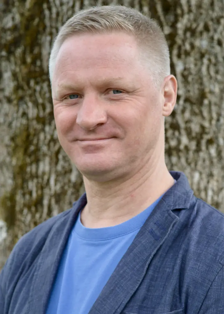 En man med ljusbrunt kortklippt hår. Han ler. På sig har han en blå T-shirt. Över den har han en mörkblå kavaj. I bakgrunden syns en trädstam med mossa. Bilden är tagen utomhus.