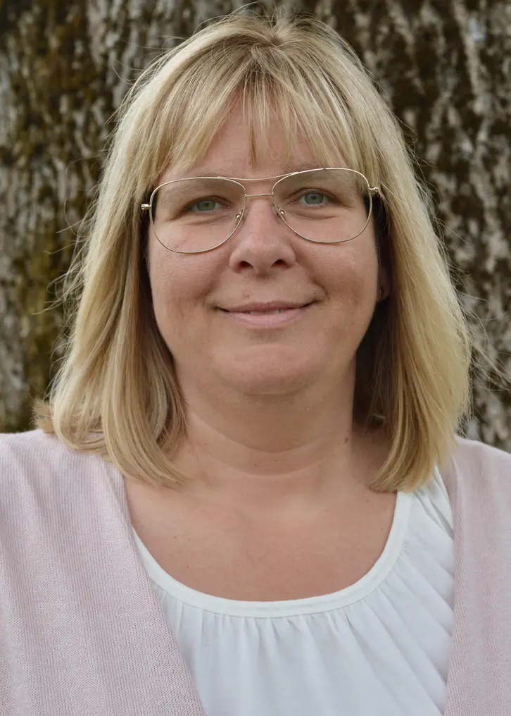 En leende kvinna med ljust axel-långt hår. Hon har pilot-formade glasögon. Hon har en vit tröja på sig. Utanpå den har hon en ljusrosa kofta. I bakgrunden syns en mossig gammal trädstam med grov bark. Bilden är tagen utomhus.