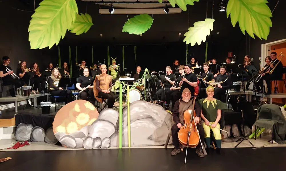 Kulturskolans Ungdomsblås sitter i bakgrunden med sina instrument. I taket hänger gröna stora löv. Framför scenen är det en kuliss som föreställer stenar. Några av lärarna, utklädda till en syrsa och en spindel sitter framför orkestern.