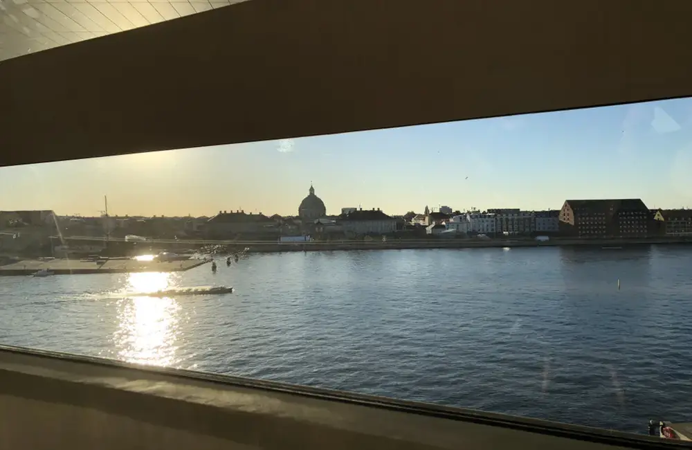 Bilden är tagen genom ett fönster . Utanför finns vatten och i bakgrunden tornar hus upp sig. Solen skiner.