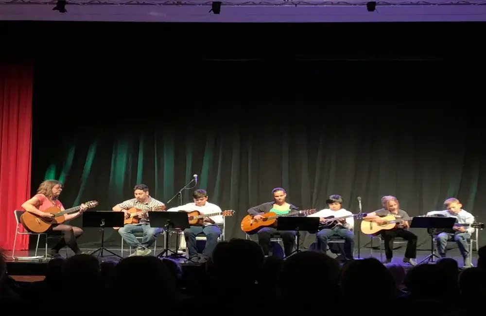 En grupp ungdomar sitter på en scen och spelar gitarr tillsammans med sin lärare. Man kan ana publikens huvud framför scenen som en svart siluett. I vänster kant syns en röd ridå.