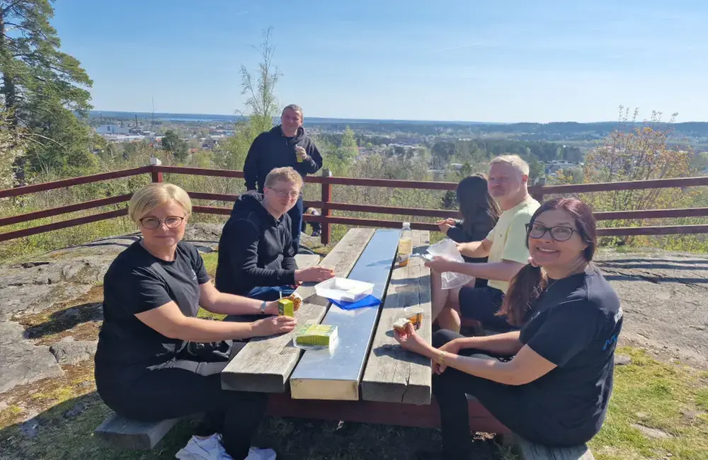 Uppe på en utsiktsplats vid ett picknickbord i trä sitter några 2 kvinnor och 3 män och fikar. Det är strålande fint väder och en blå himmel. I bakgrunden ser man ett rött staket.