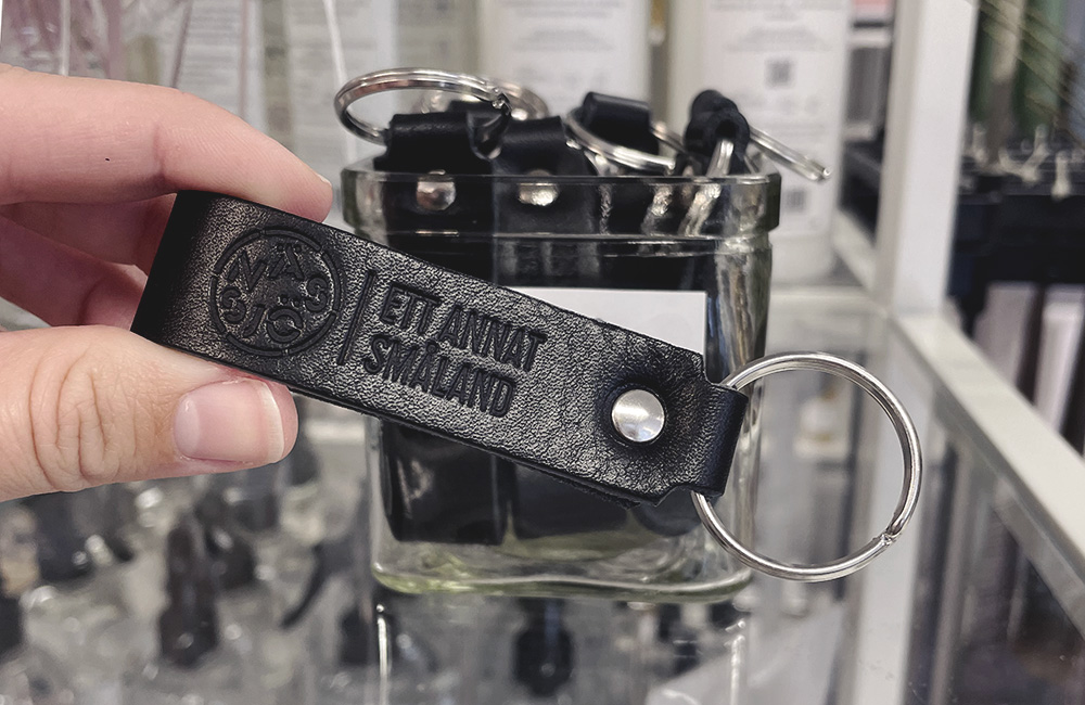 En hand håller fram en nyckelring i svart läder med texten "Ett annat Småland" samt platsvarumärkets logotyp. I bakgrunden ser man en hel skål med likadana nyckelringar.