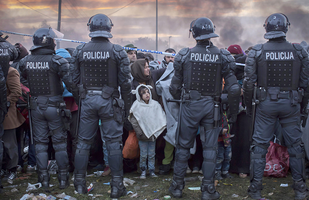Ett foto av en folkmassa med flyktingar med en rad poliser som står framför dem. I centrum av bilden står en liten flicka och tittar upp på poliserna.