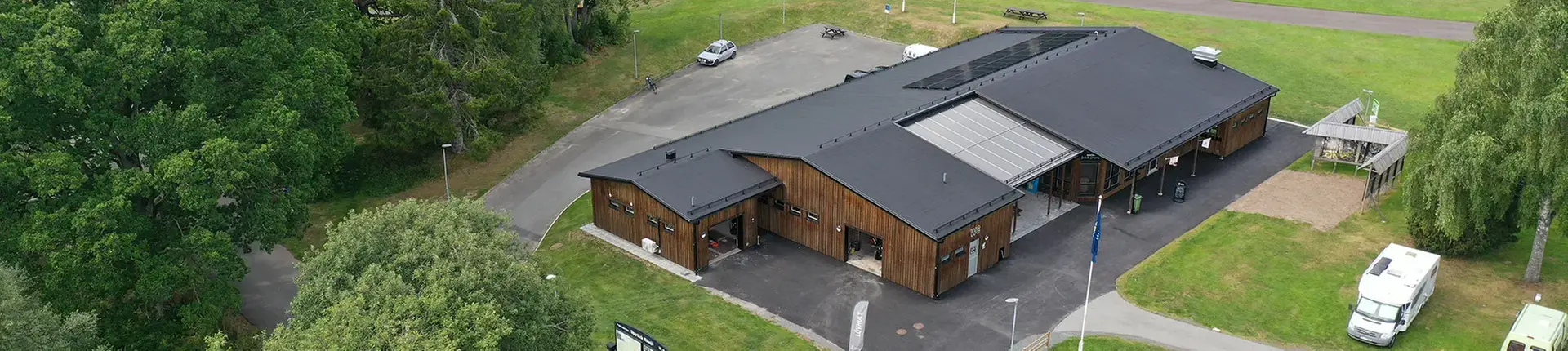 Drönarbild på träbyggnad med brun panel och svart tak. Till vänster finns en skog och runt om byggnaden gräsmatta med två husbilar på.